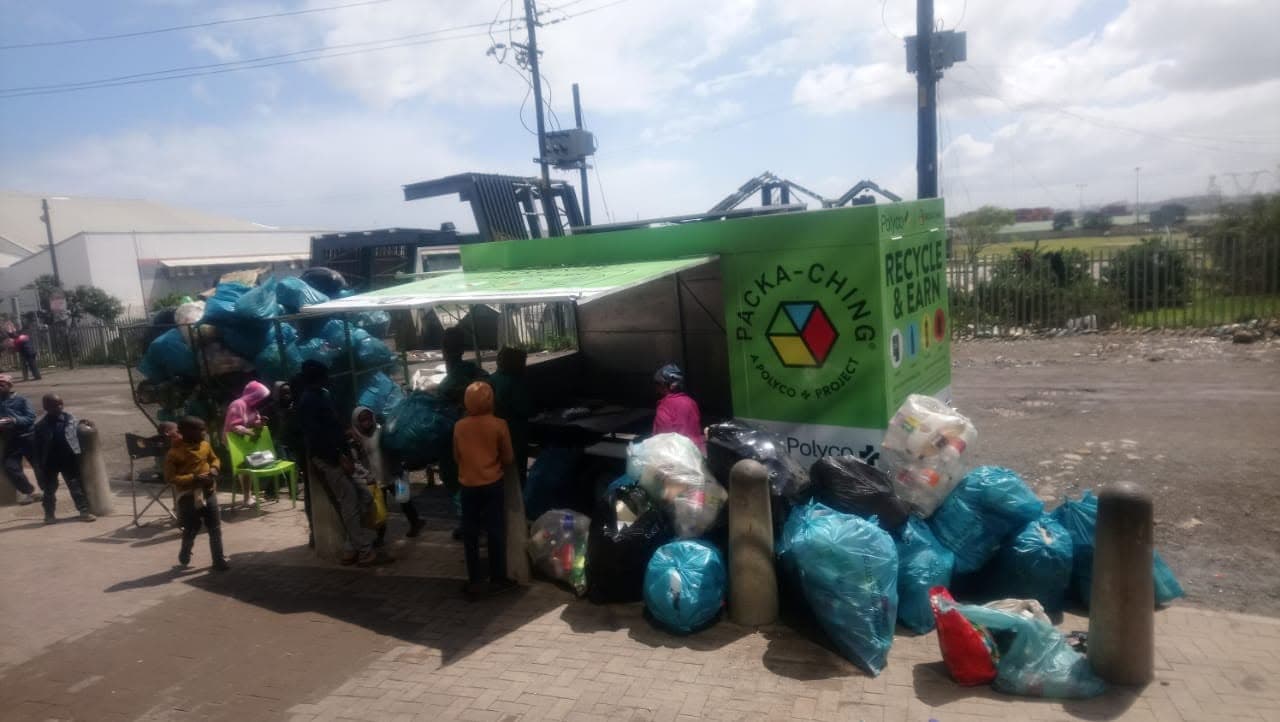 Community members gathered around a Packa-Ching mobile buyback unit surrounded by bags of recyclables