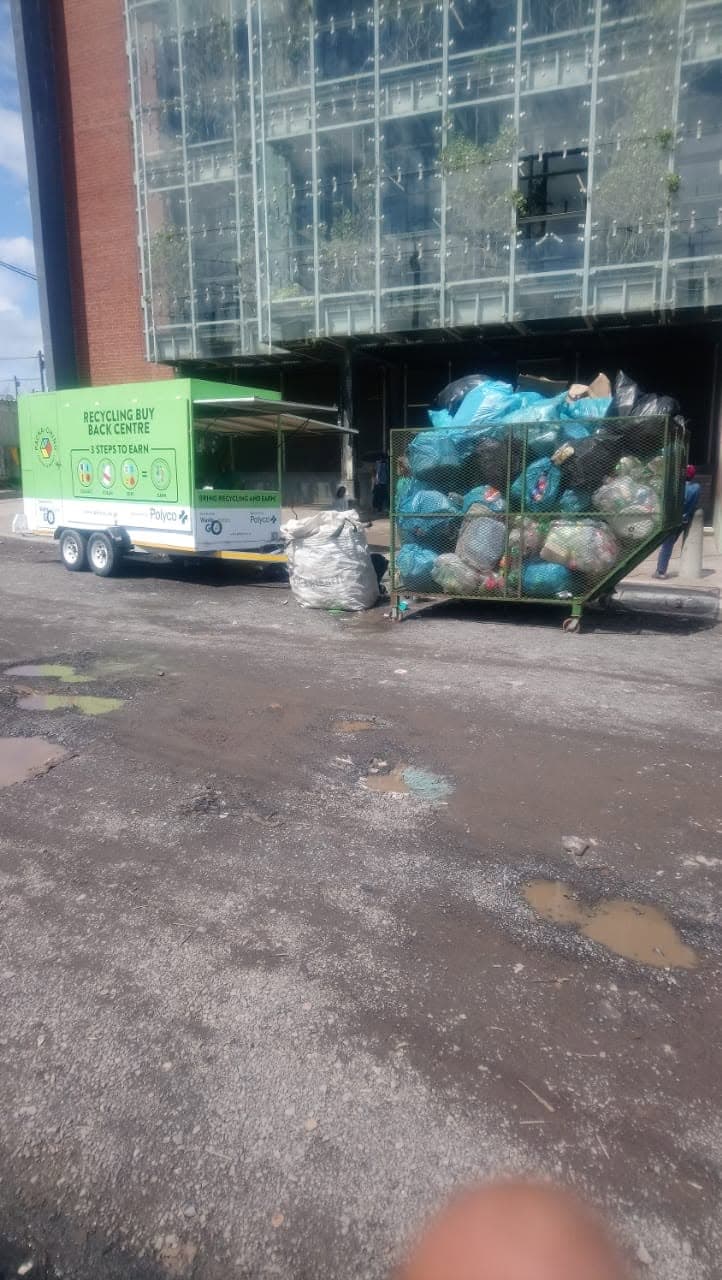 A green ‘Recycling Buy Back Centre — 3 steps to earn’ trailer next to a cage trailer full of sorted recyclables