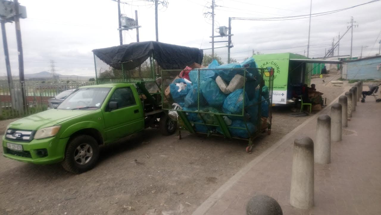 A green bakkie and cage trailer filled with blue bags of recyclables beside a Packa-Ching unit in a Cape Town township